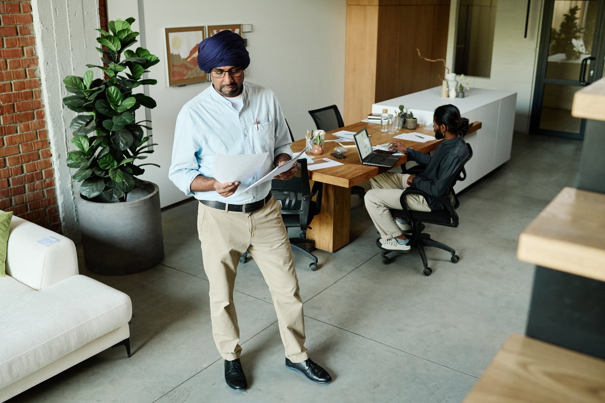 Indian workers in modern office, older one standing in center of office and reading report while younger one sitting at table and making presentation