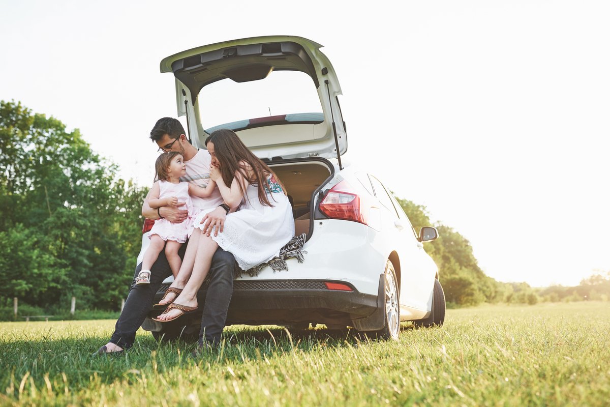 Pretty young married couple and their daughter are resting in the nature. The mother father and little girl are sitting on open car boot.