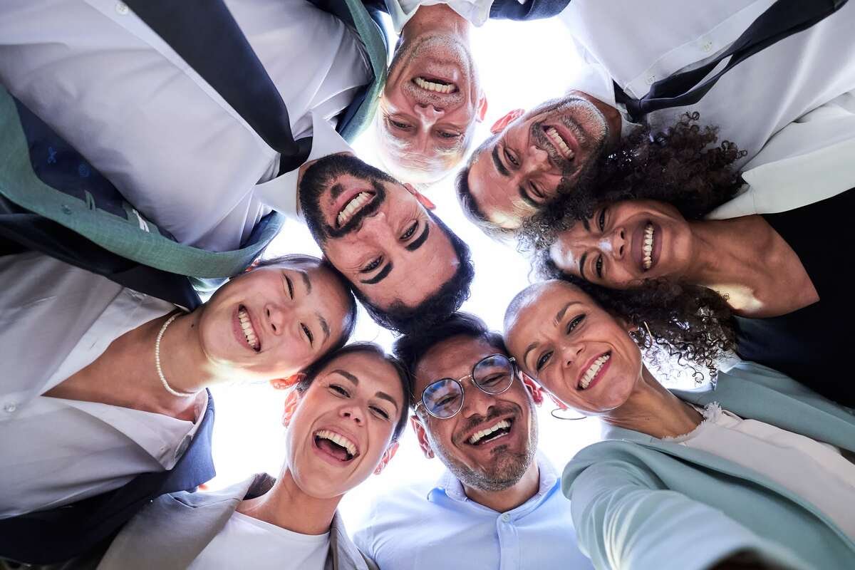 Multiethnic group of work colleagues embraced in circle, face down taking a selfie smiling and looking at the camera. Vertical image, POV business people doing team building.